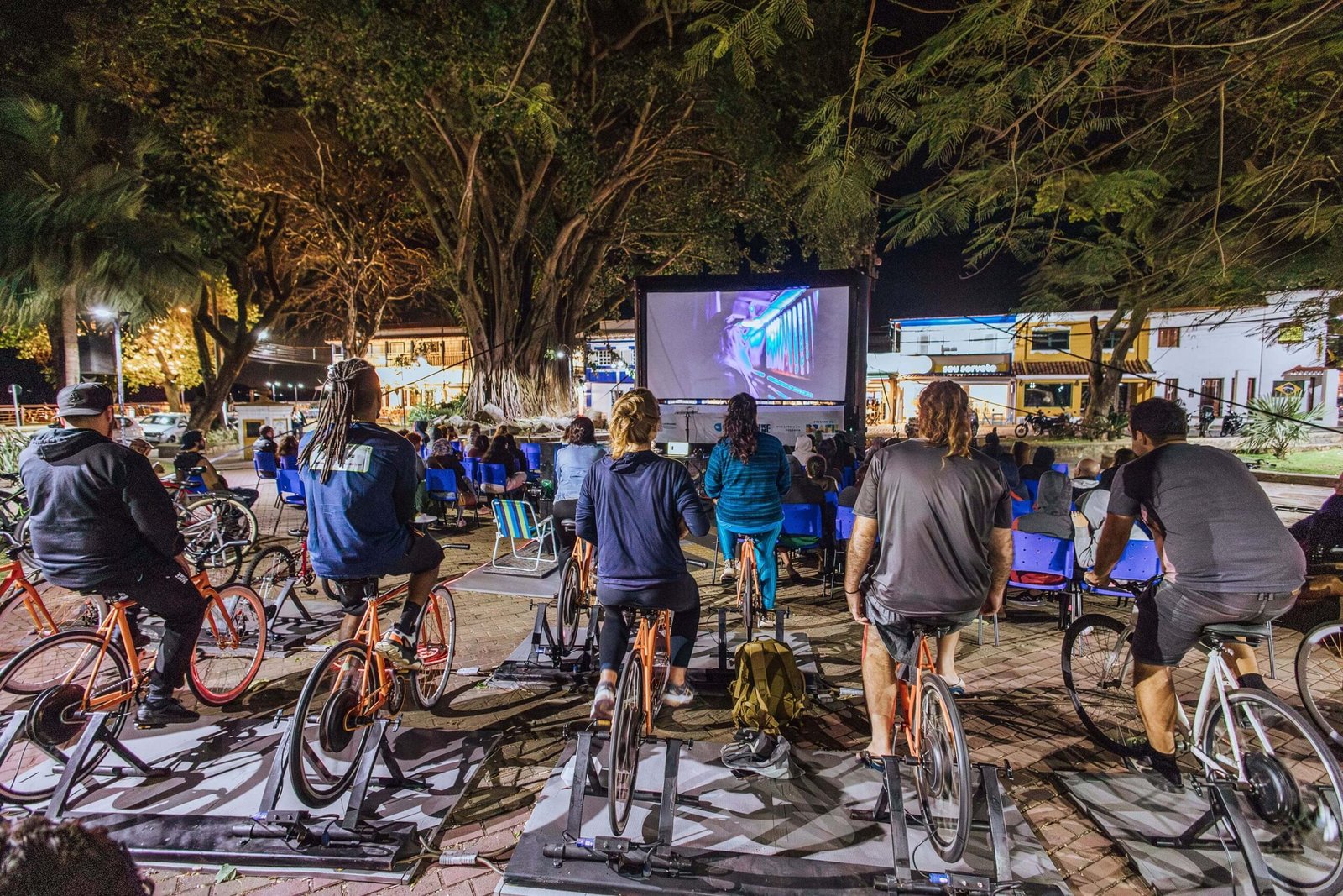 Fotografia em plano geral, retratando um evento de cinema ao ar livre em uma pra&ccedil;a p&uacute;blica. Em primeiro plano, pessoas de costas pedalam bicicletas laranjas fixas enquanto assistem a um filme na tela grande &agrave; frente. Ao fundo, &aacute;rvores grandes e casas iluminadas.